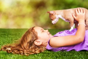 Cute little girl laying on grass outdoors with puppy on her chest looking down at her